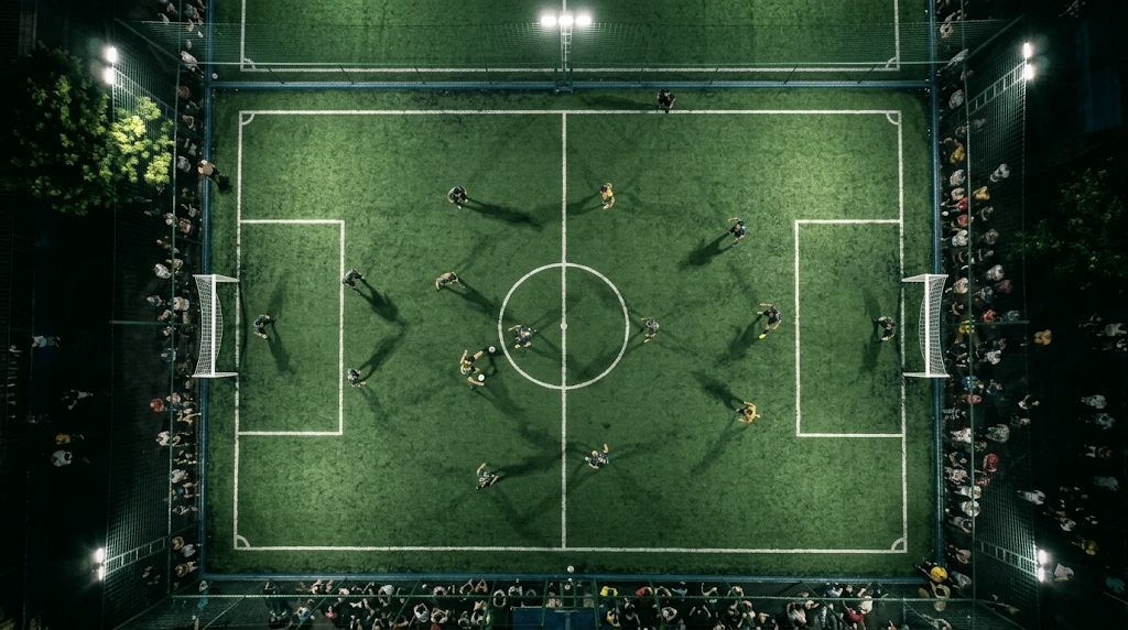 Vista aérea de um campo society iluminado à noite, com jogadores em campo